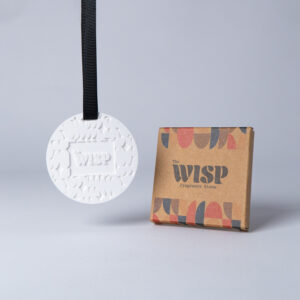 The Wisp Fragrance Stone — embossed ceramic scent stone on a black ribbon beside its kraft box